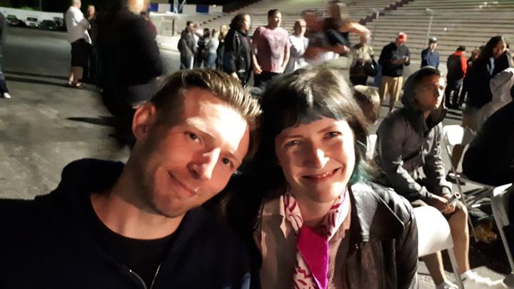 Man and woman smiling with people conversing at an outdoor event in the background. 