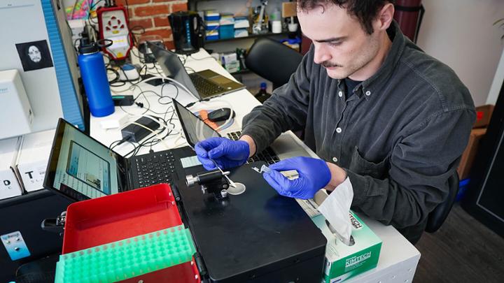 Man with a moustache and wearing surgical gloves tests an unknown substance using device hooked up to a laptop computer. 