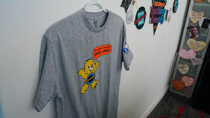 Grey T-shirt hanging on a hanger depicting a cartoon bear with a dialogue bubble that reads "drug checking saves lives"