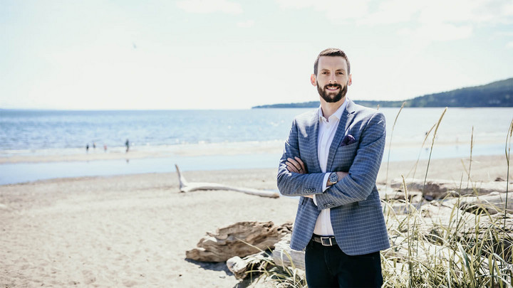 Man with beard in blue sports jacket standing cross-armed in front of seaside beach.