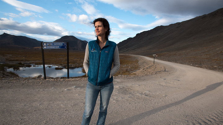 Woman in a vest standing outside in front of a road sign and hills in the background.