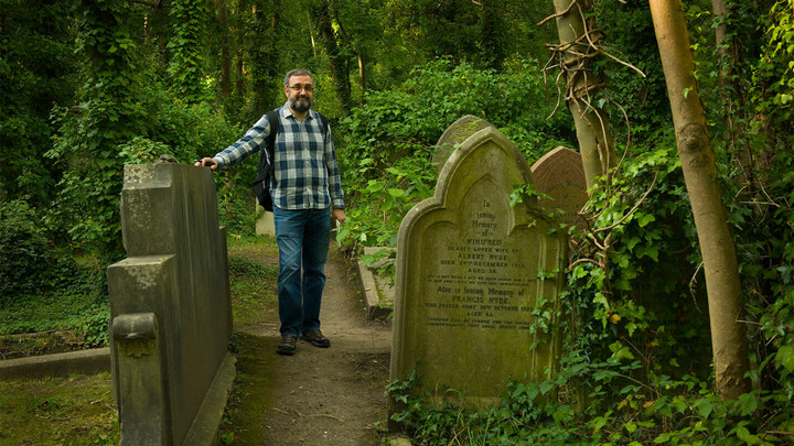 Man with beard and glasses standing next to grave stones in a forested area.. 