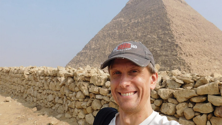 Man in a baseball hat smiling while standing in front of an Egyptian pyramid.