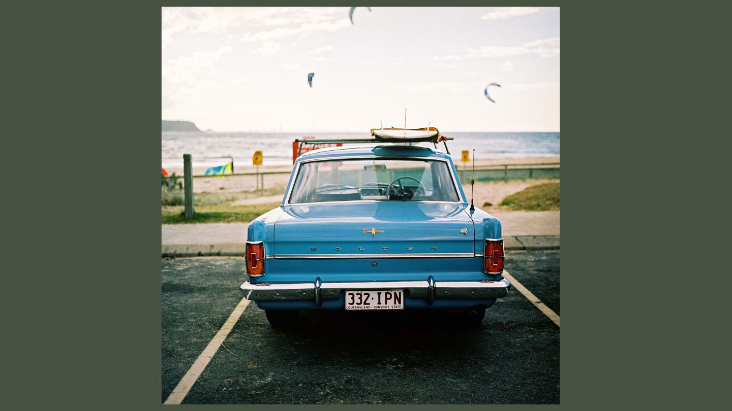 Vintage car with surf board strapped to the roof parked in front of a beach.