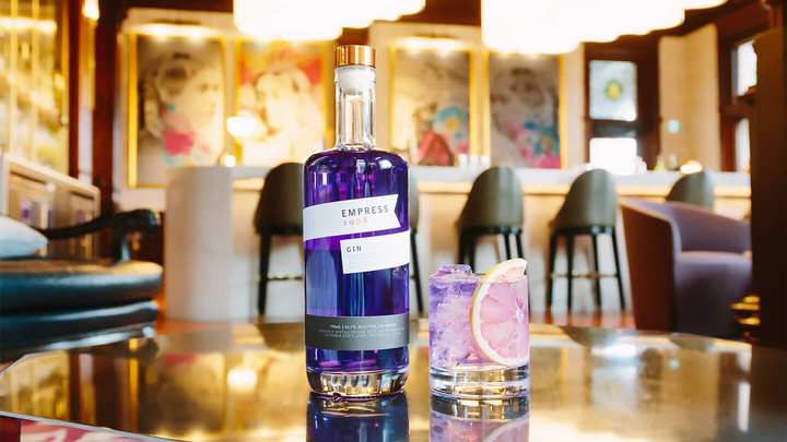 Bottle of purple coloured Empress Gin beside a purple hued cocktail. 