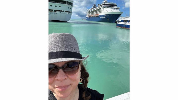 Woman wearing a hat and sunglasses in front of two cruise ships at dock.