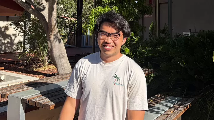 A person with glasses and a T-shirt smiling while sitting at an outdoor table surrounded by trees.