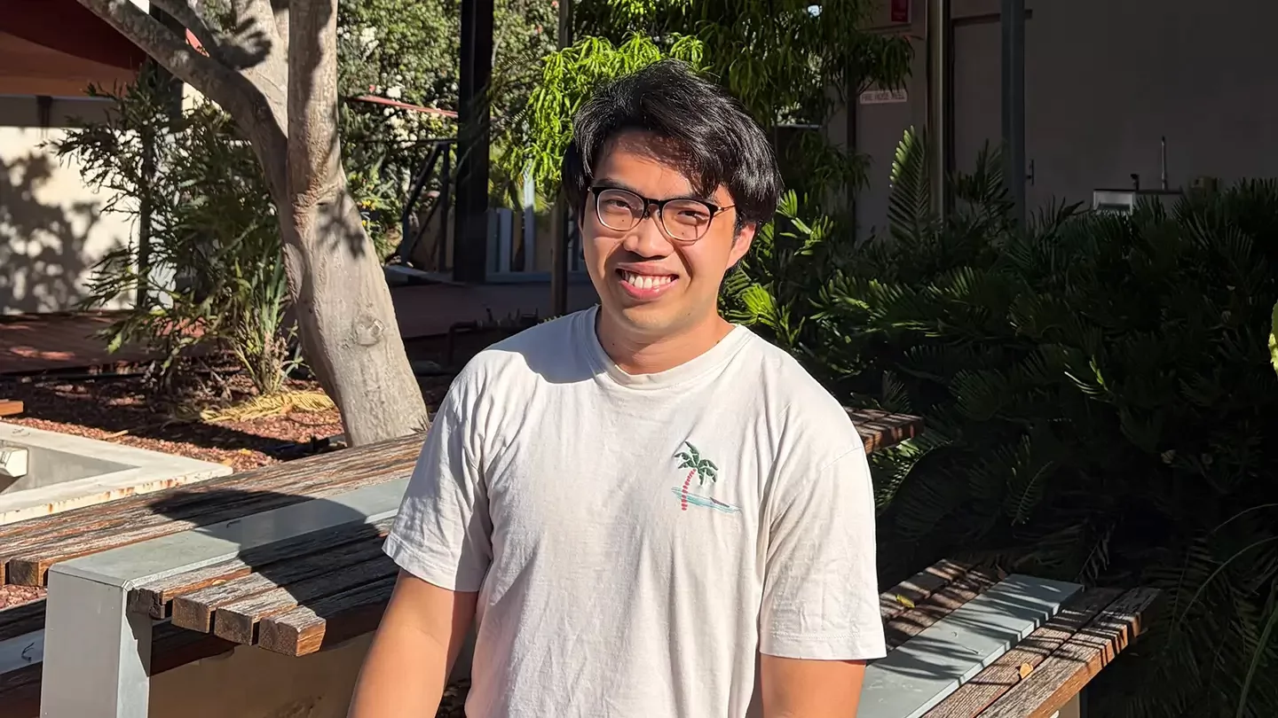 A person with glasses and a T-shirt smiling while sitting at an outdoor table surrounded by trees.