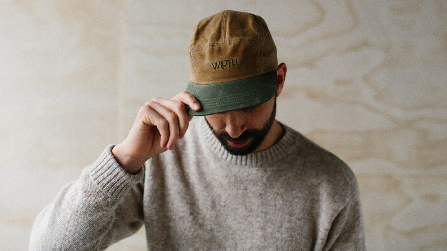 Profile of a man with a grey sweater and brown-green baseball cap using the hat’s brim to hide his eyes from the camera. 