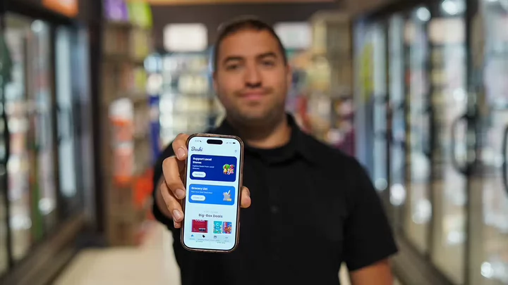 Yashar Samin holding up a smartphone displaying the Baski app while standing in the frozen foods aisle of a grocery store. 