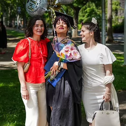 A woman smiling in a graduation gown and hat holding flowers and balloons with a woman on each side of her also smiling.