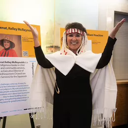 A woman wearing an Indigenous blanket smiling and raising her arms in the air.