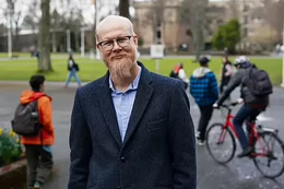 A man with horn-rimmed glasses and a beard smiling while standing on a university campus lawn.