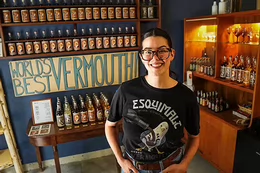 Woman with glasses and shirt that reads Esquimalt Vermouth & Aperitifs standing in front of a shelf of bottles and a handmade sign that reads World's Best Vermouth. 