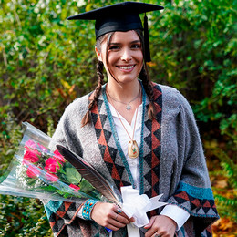 Graduating student smiling and wearing cap and ceremonial blanket while holding flowers and a feather.