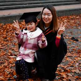 A woman smiling in a graduation gown with her toddler daughter who's wearing a graduation cap.