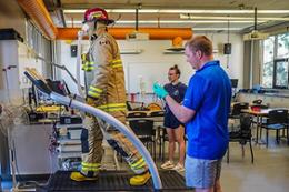 Man and woman monitor a firefighter in full gear walking on a treadmill. 