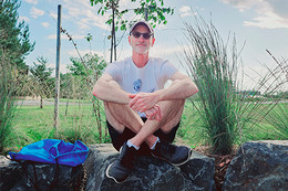 Person sitting crossed legged outdoors, wearing shorts, T-shirt, running shoes, sunglasses and baseball cap.
