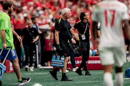 A man with white hair wearing a black T-shirt and shorts walks on a sports field during a game carrying bottles of Powerade..