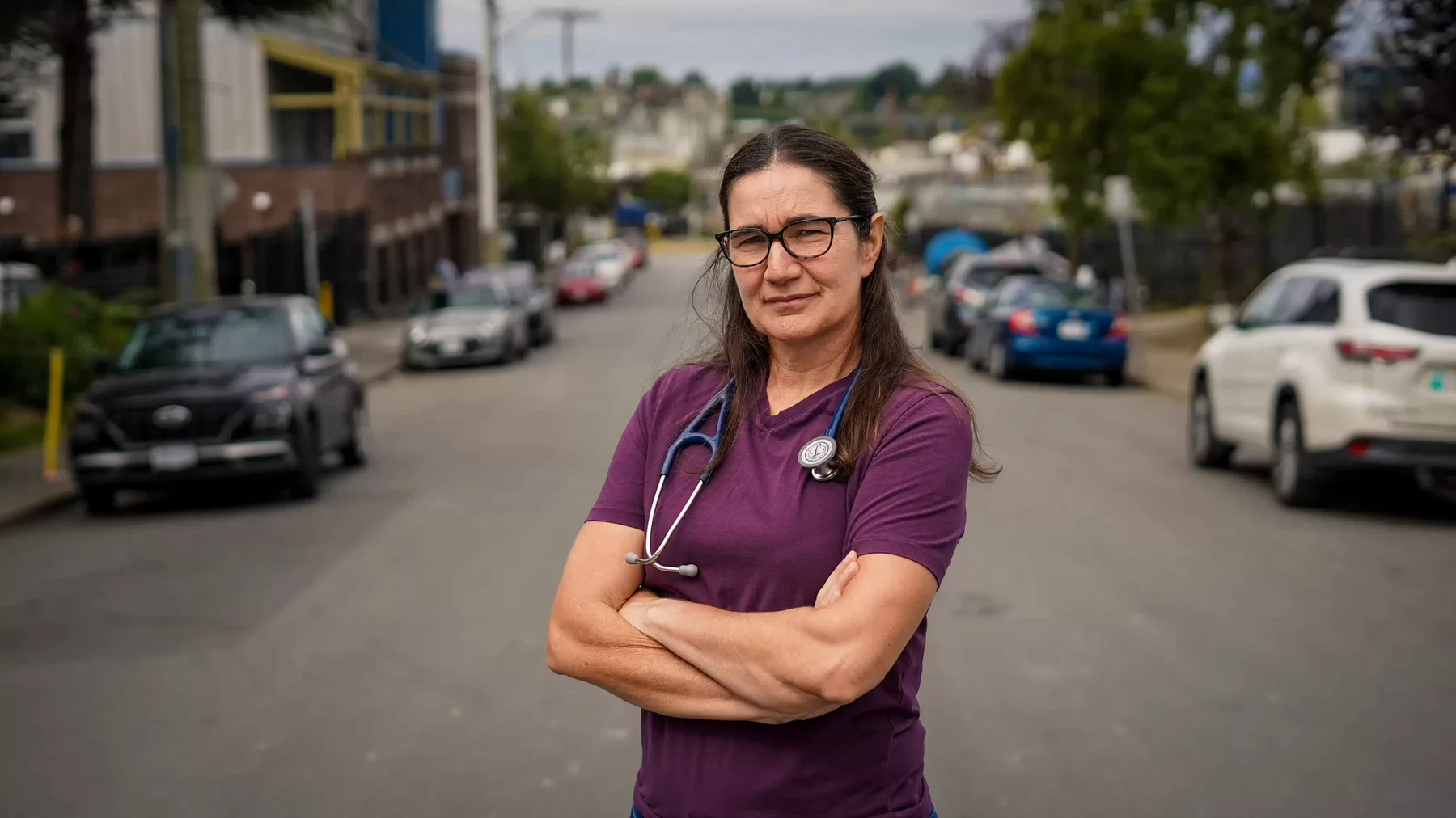 A woman with glasses and stethoscope around her neck standing with her arms crossed in the middle of a road.