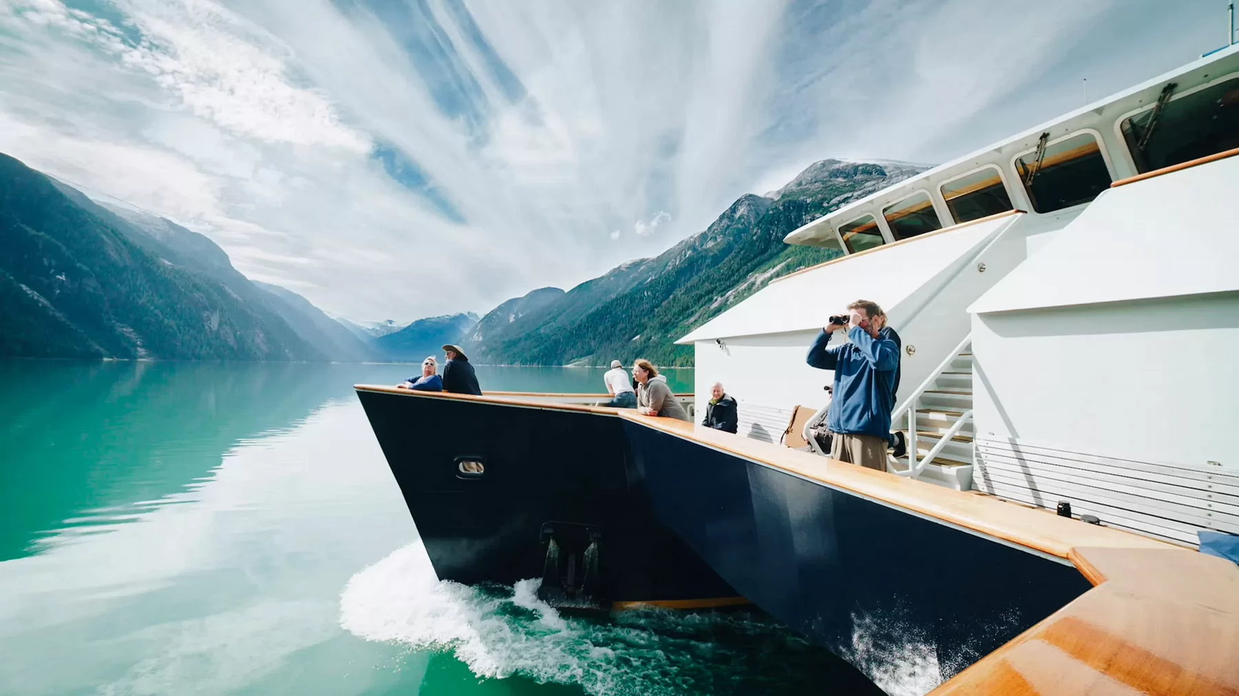 People gathered on a deck of a boat taking in the views of an inlet surrounded by mountains. 