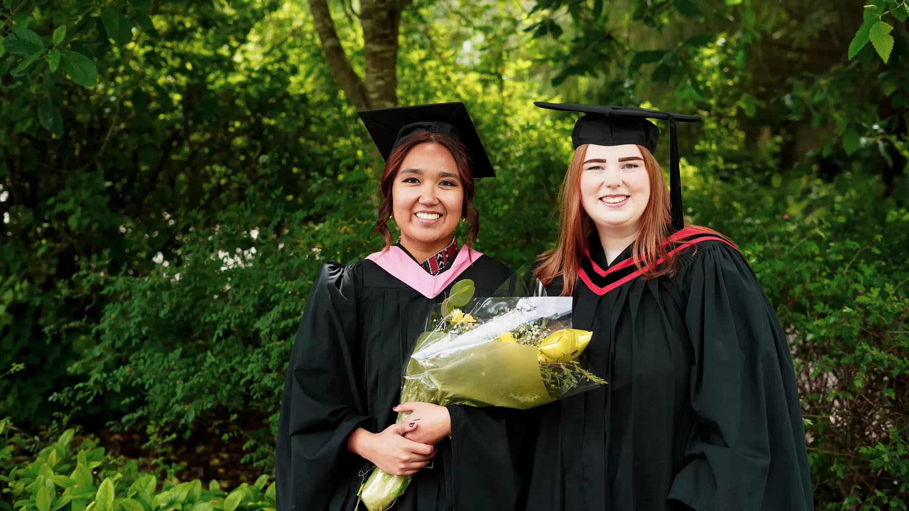 Two university graduates wearing caps and gowns smiling while standing in front of shrubbery. 