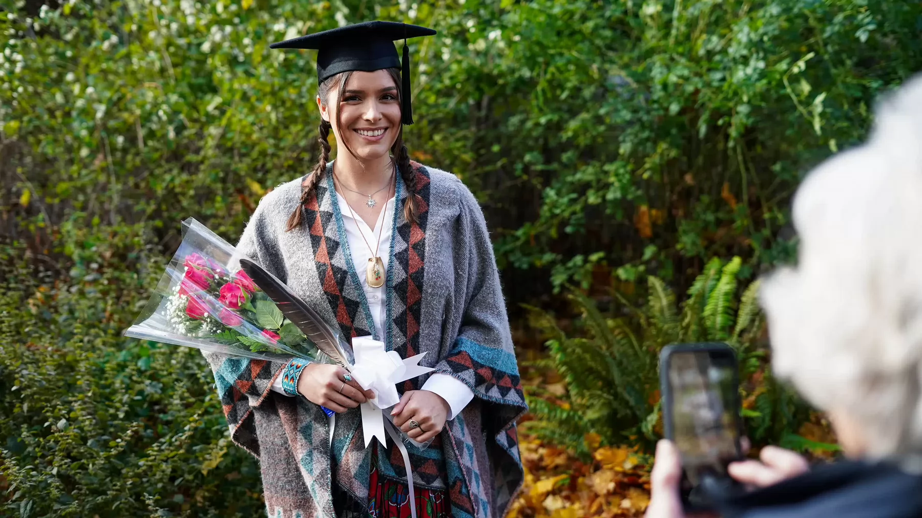 Woman wrapped in a Indigenous blanket, wearing graduation cap and smiling for camera while holding a feather and flowers.