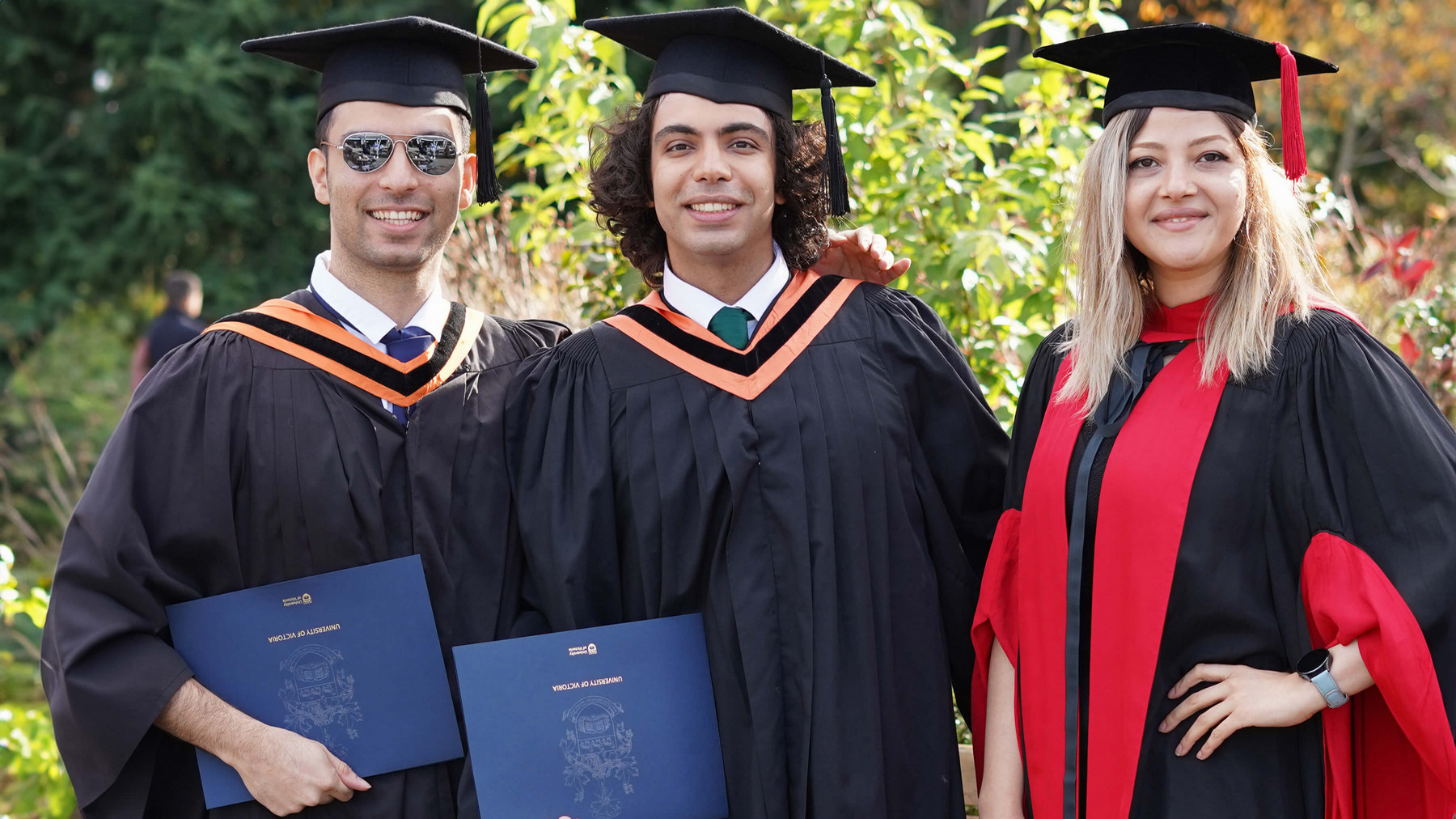 Three graduating students standing outdoors side by side smiling in graduation robes and hats holding diplomas.