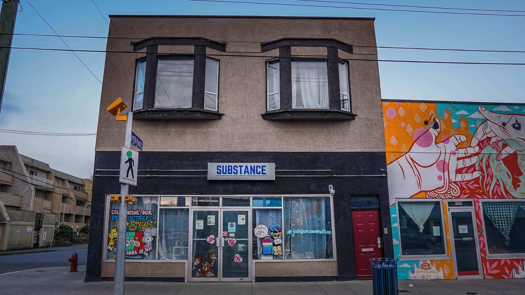 Storefront of a two storey grey stucco building with drawings of cartoon bears on the windows and a neighbourhing building decorated with mural art of two outreached hands touching.