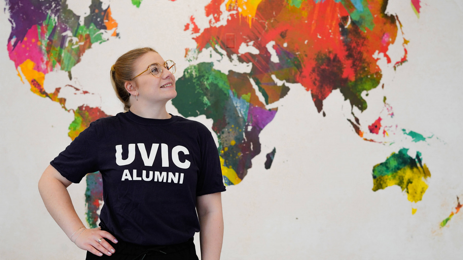 Woman wearing a T-shirt that says UVic Alumni standing in front of a map of the world.