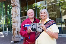 A man and a woman holding up black and white photos of their younger selves in front of UVic’s First Peoples House.