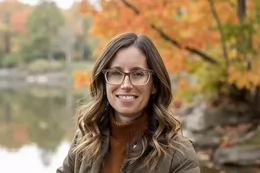 A woman with glasses smiling in front of a tree-lined body of water.