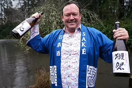 A man in a Japanese robe that says Sake on the lapel holding up two bottles of sake.
