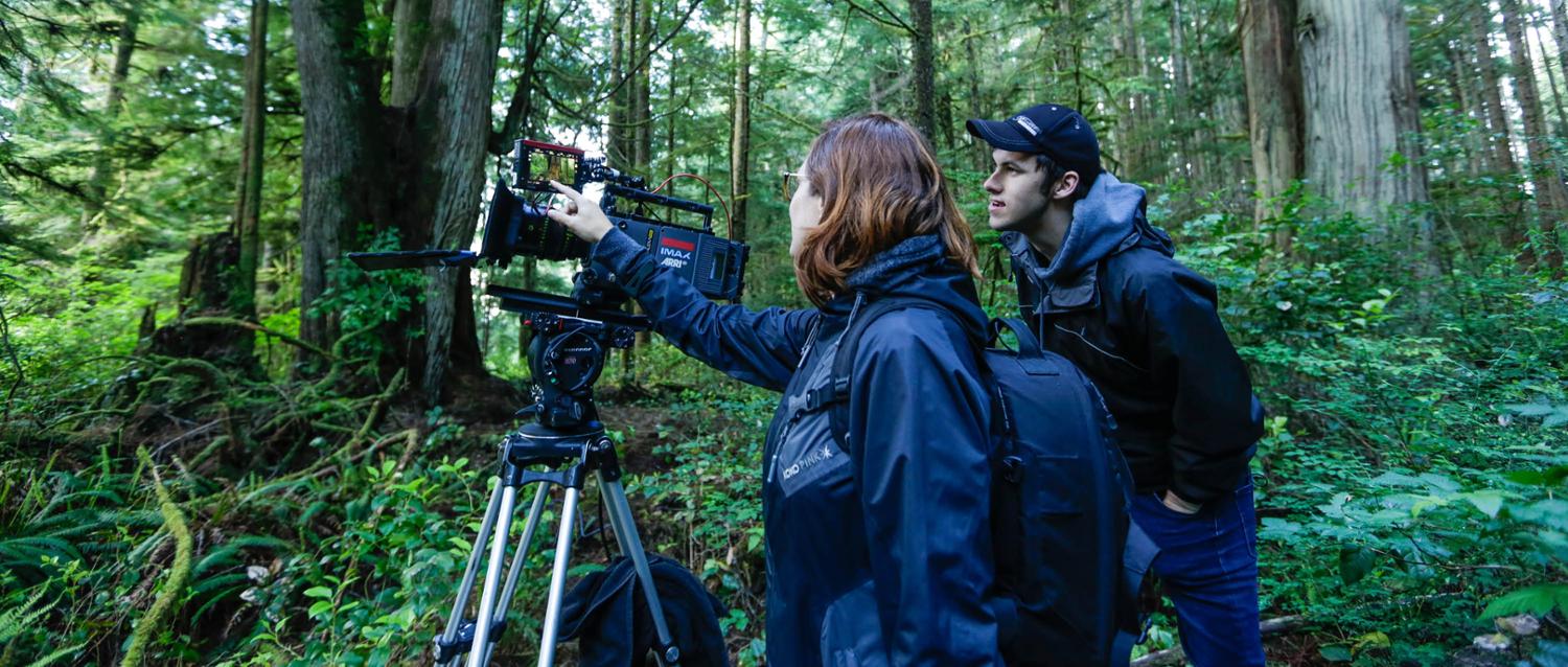 Kelly Richardson and student hold a video camera surrounded by forest.