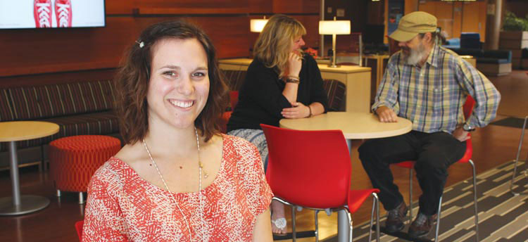 A student facilitator sits facing the camera. A group of students talk with each other in the background.