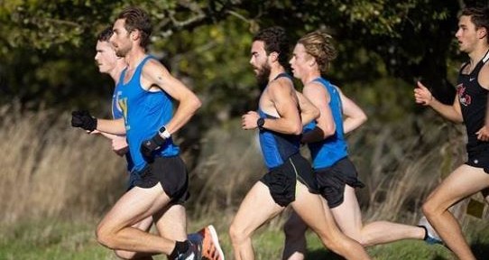 Five men running, photographed from the side. There is a forest in the background. 