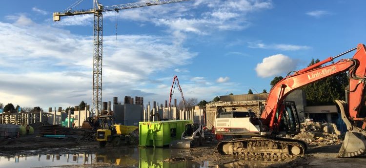 Construction site with an excavator in the right side of the photo, a shipping container in the centre, building scraps all around, and a crane in the distance.