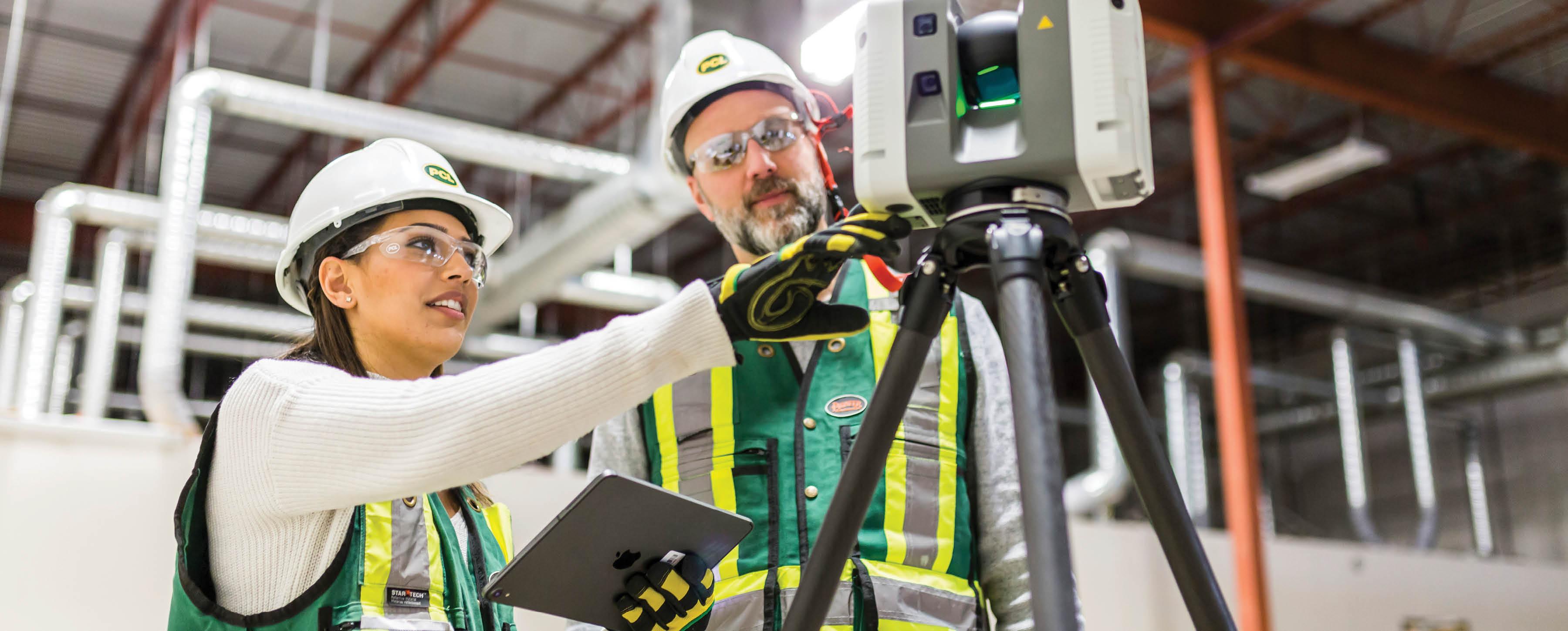 An older man and a younger woman are dressed in high-visibility vests, white hard hats, and goggles. They are discussing a complicated technical problem.