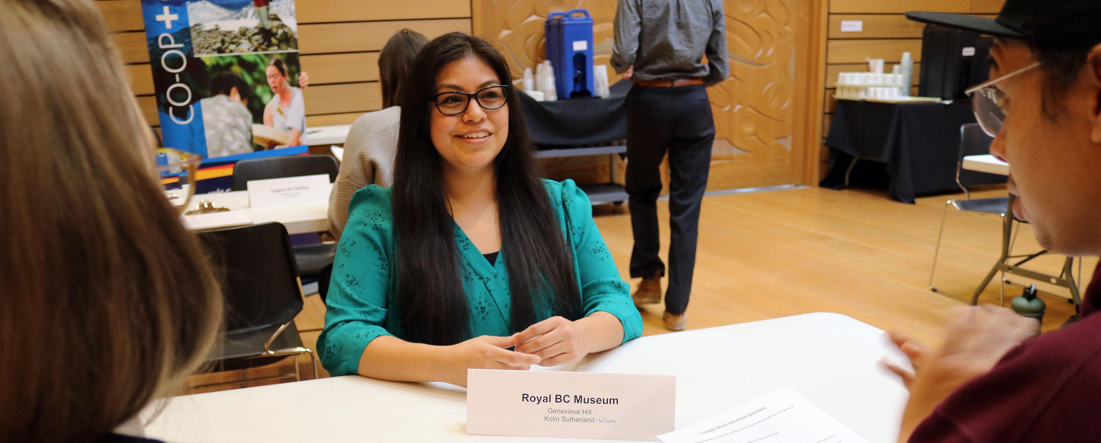 An Indigenous student sits at a table facing the camera. There are two people on the other side of the table looking at the student. This is an interview.