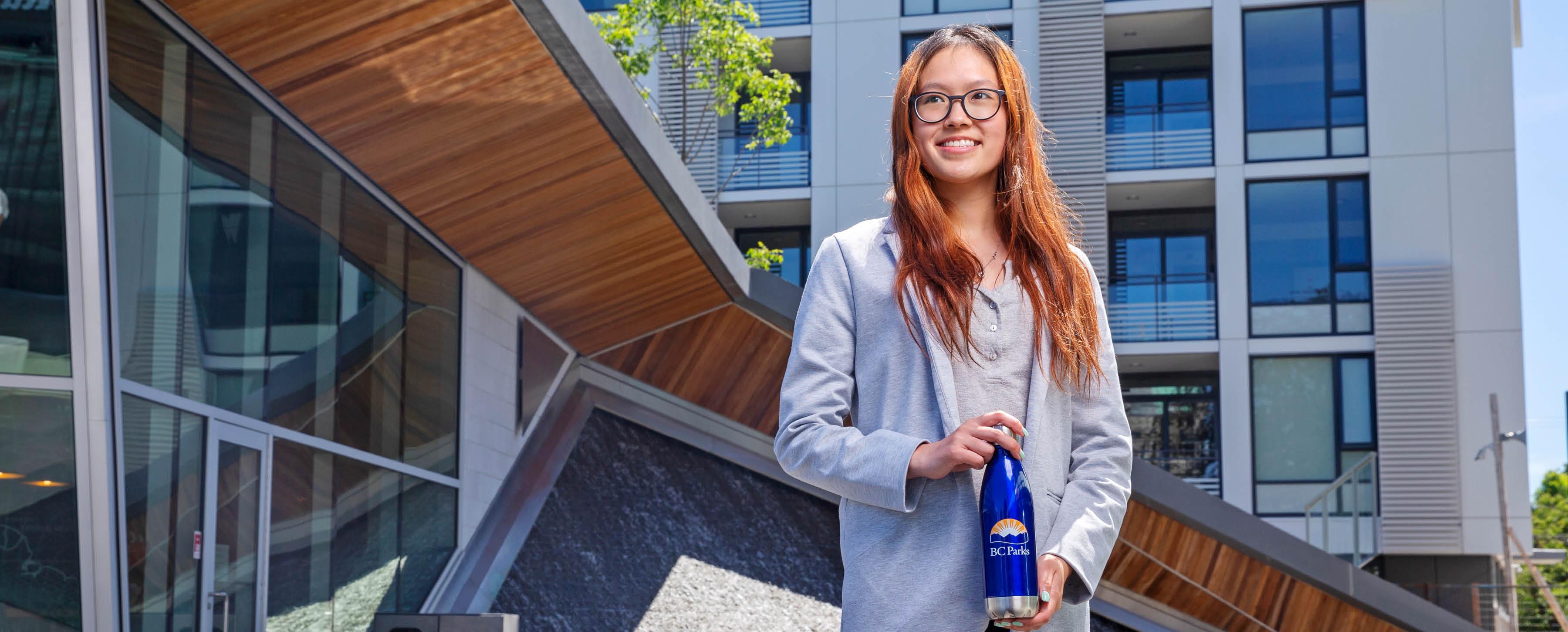 A UVic student is standing in front of a modern-looking glass and steel building. She is dressed professionally in a blazer and is smiling towards the camera.