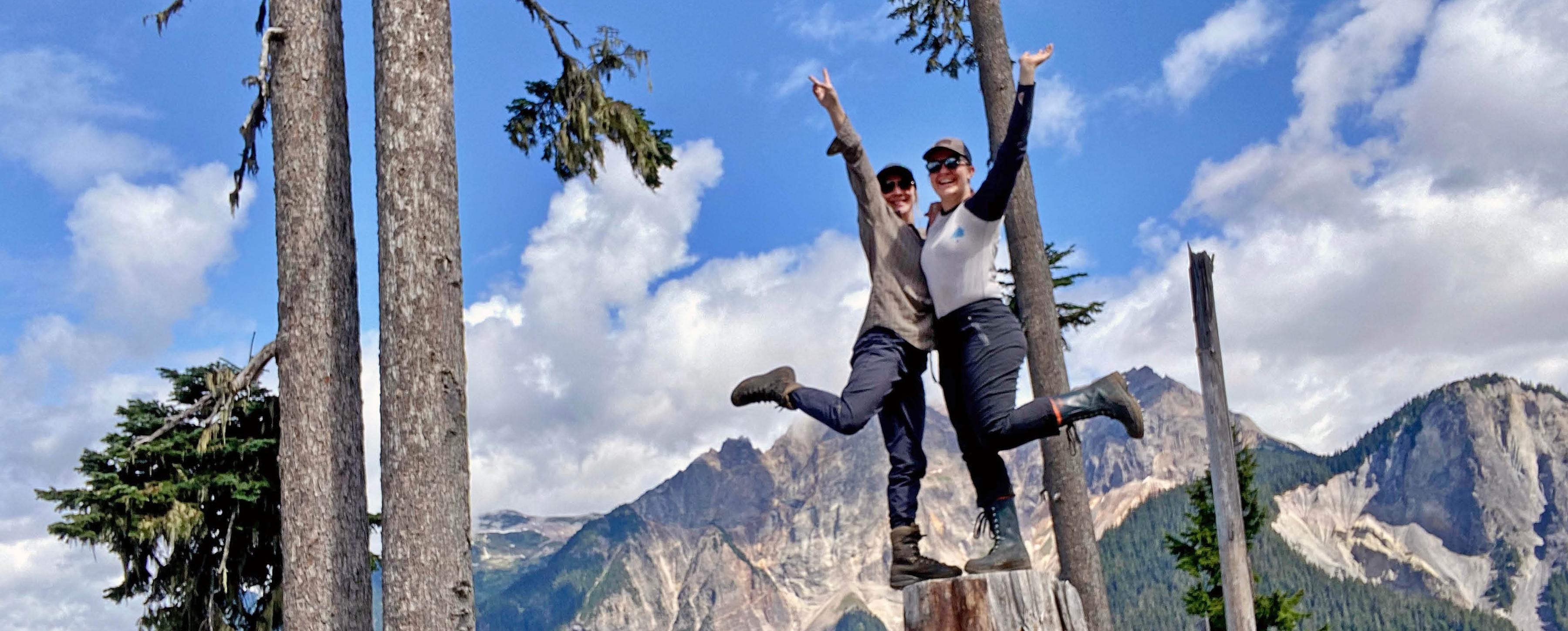 Two students are posing happily on a tree stump, with mountains and forest in the background.
