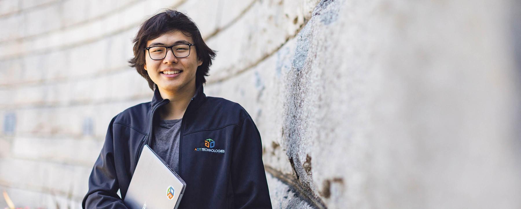A young student of Asian descent is standing outside, wearing a black jacket and glasses and holding a clipboard, smiling at the camera.