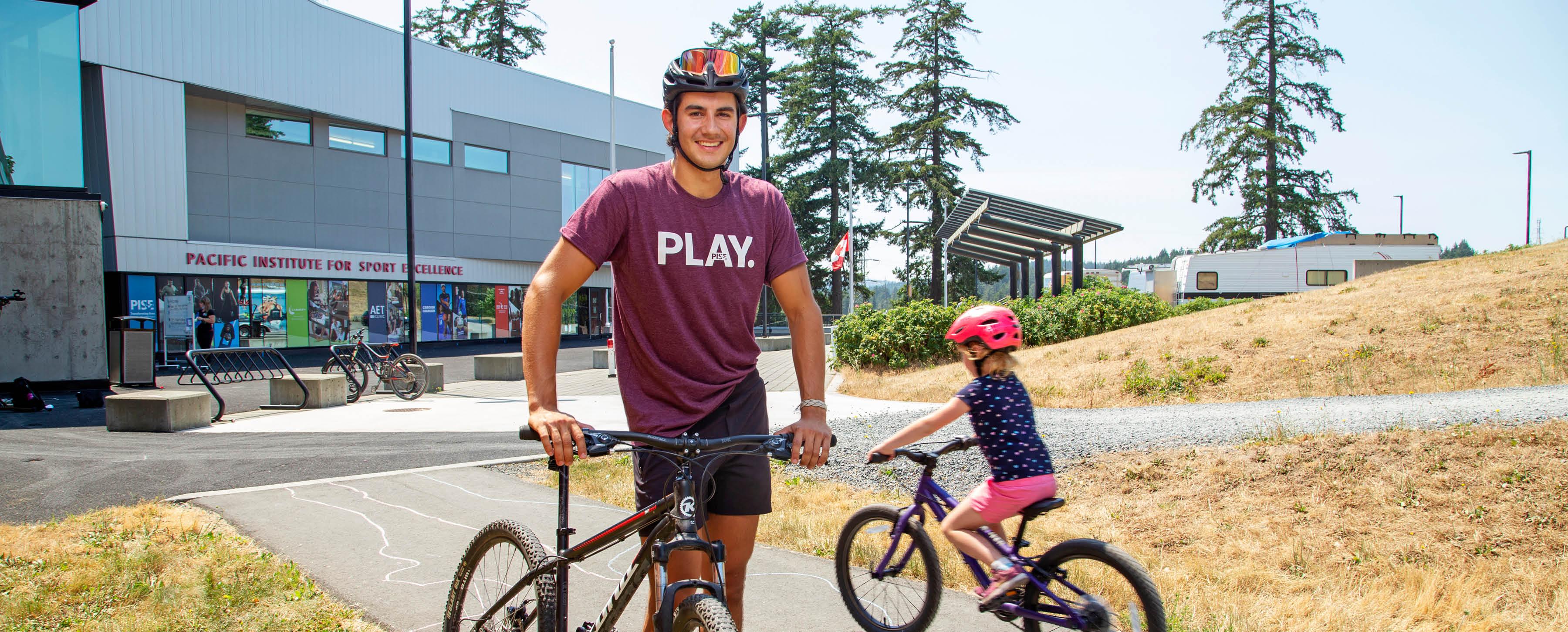 A man wearing shorts, a burgundy t-shirt that says "Play," and a black bike helmet with goggles is posing with a bicycle in front of a field with a building and RVs in the background. He is smiling at the camera.