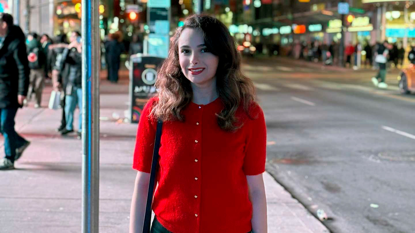 A student wearing a bright red sweater stands on a sidewalk in front of a busy street at night.