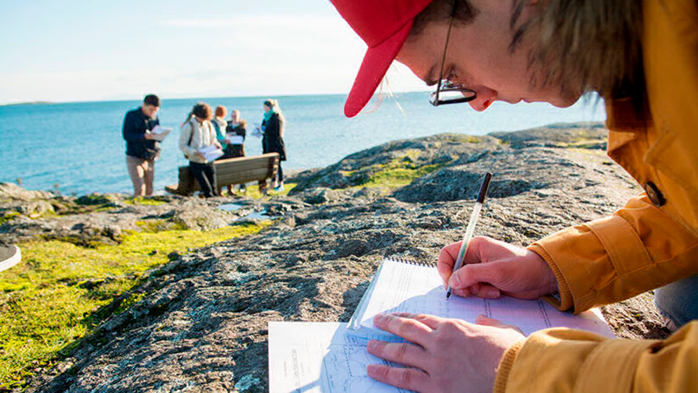 A student writes on a notepad on a rocky outcrop, while other students can be seen in the distance.