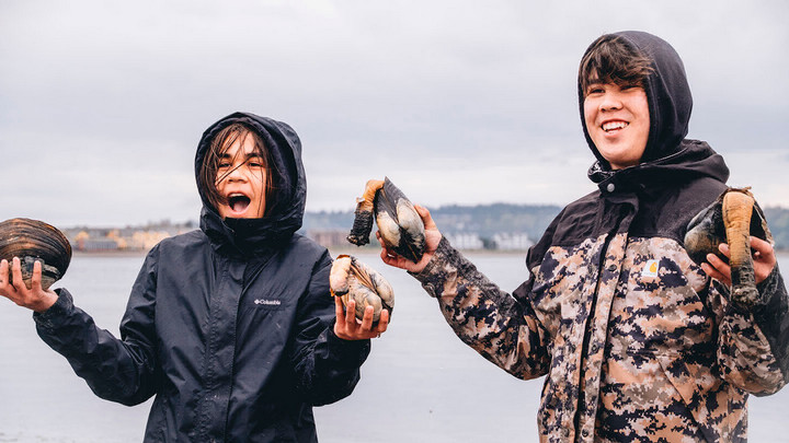 Two young people stand on a shoreline holding shells.