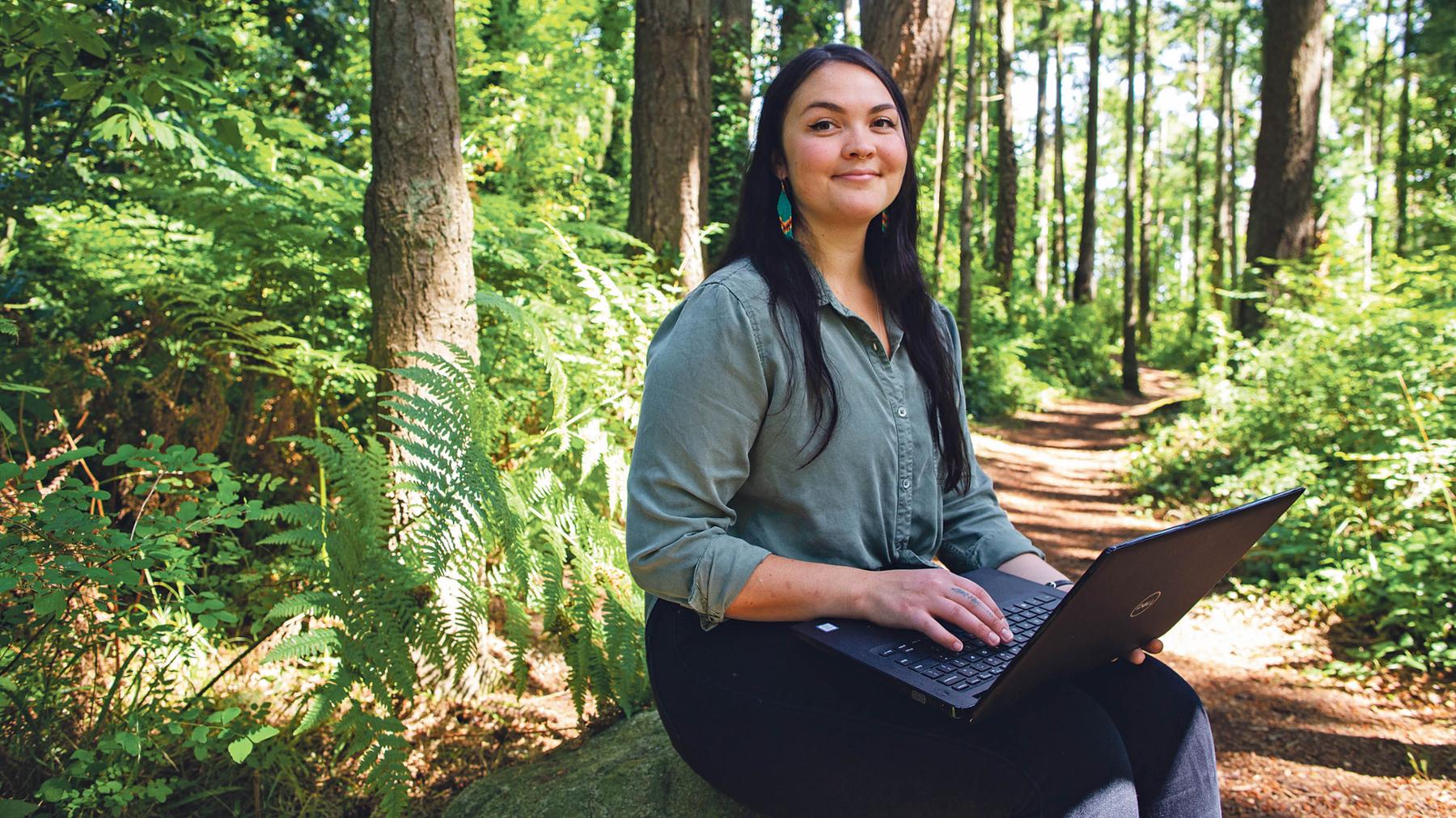 A woman with long hair and a blue shirt sits on a rock in the forest. She is using a laptop.
