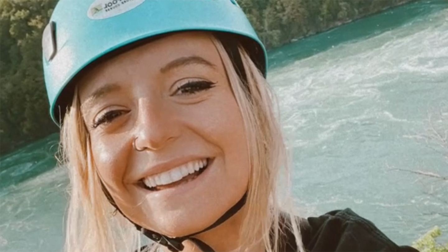 A female student wearing a rafting helmet stands in front of a river.