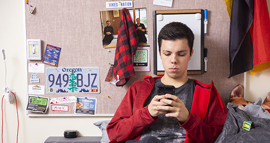 Student sitting on his dorm bed looking at his phone