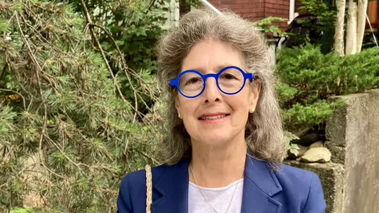 Eloise Spitzer, wearing a blue blazer, stands outdoors with greenery and part of a building in the background.